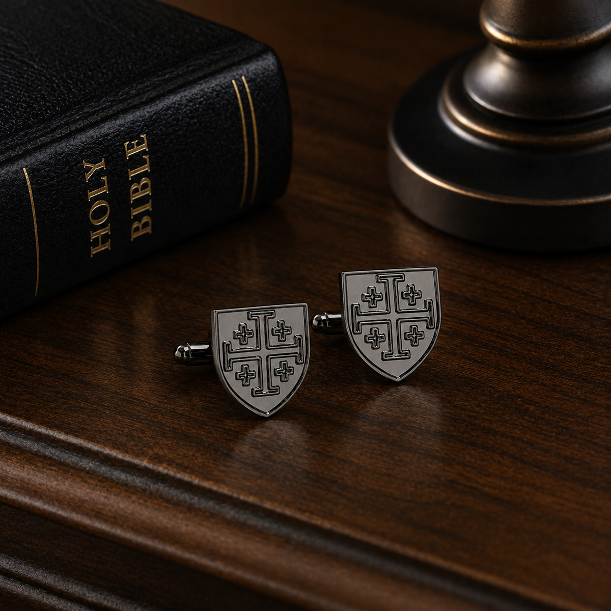 A pair of gunmetal-toned cufflinks shaped like medieval shields with black cross motifs, displayed on a dark wooden surface next to a black Holy Bible and a brass object.
