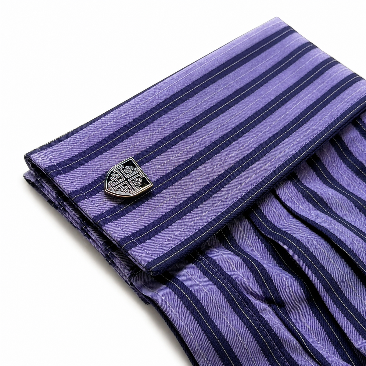 A folded purple and navy striped cufflinks shirt with a gunmetal-toned shield-shaped cufflink, shown on a clean white background.