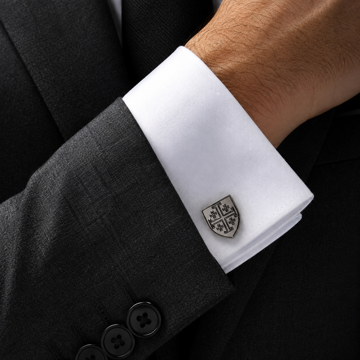A close-up of a man's wrist wearing a white dress shirt cuff with a silver-toned shield-shaped cufflink, paired with a dark gray suit jacket. Gunmetal Jerusalem Cross shield cufflinks for men
