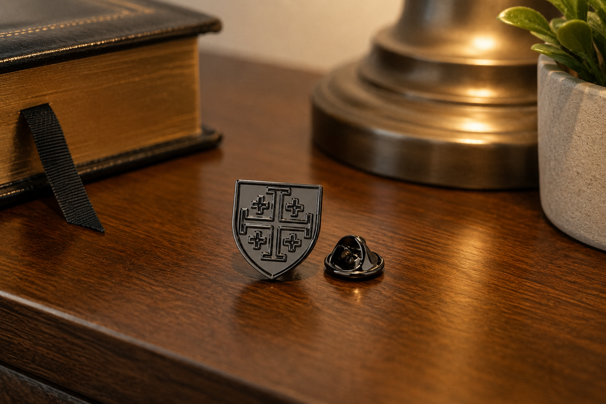 A small black enamel shield-shaped pin with a cross motif sits on a polished wooden table next to its metal backing, with a book, lamp, and plant in the softly lit background.
