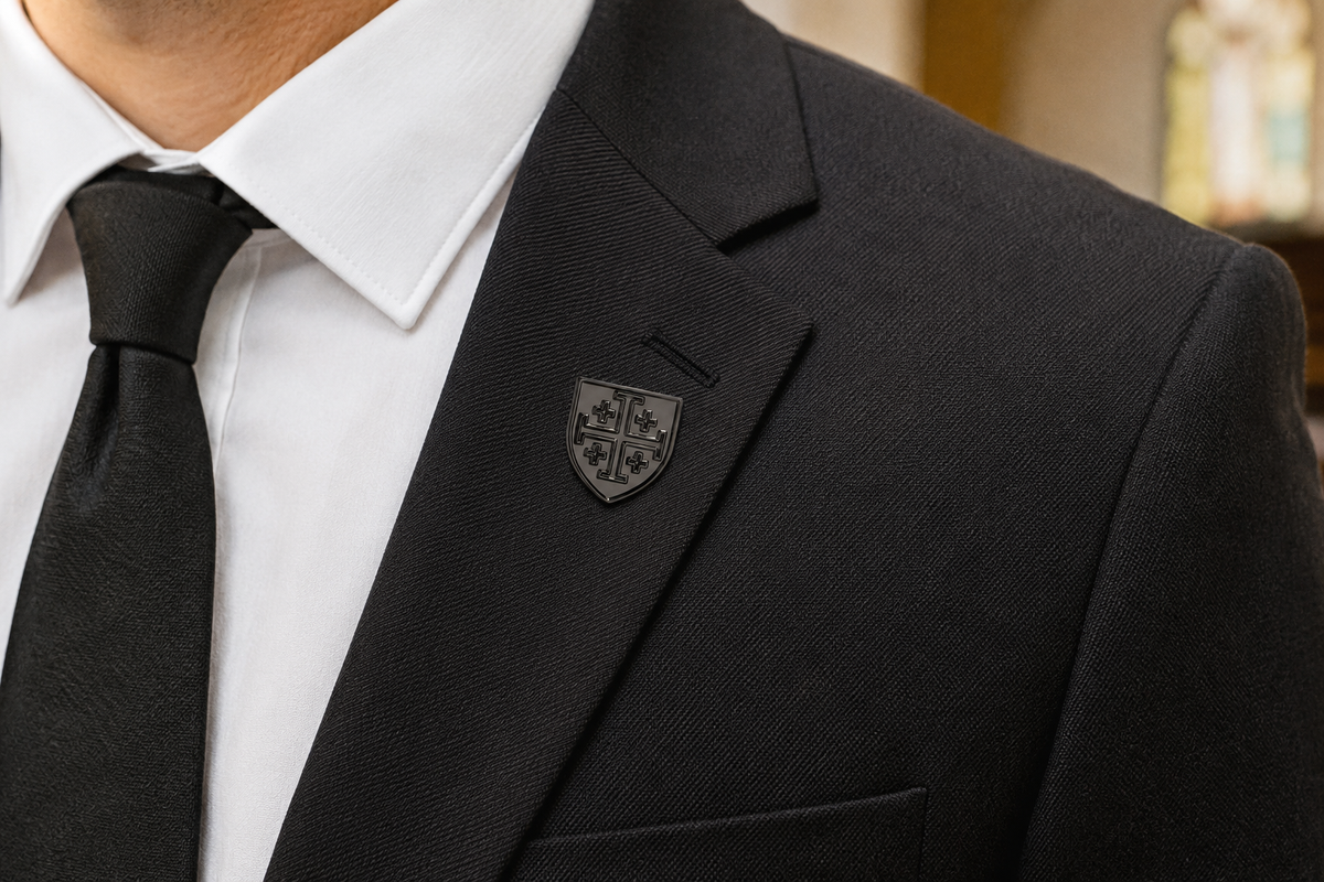 Close-up of a man wearing a black textured suit jacket, white dress shirt, black tie, and a black shield-shaped lapel pin with a Gunmetal Jerusalem Cross shield lapel pin for men with clutch back