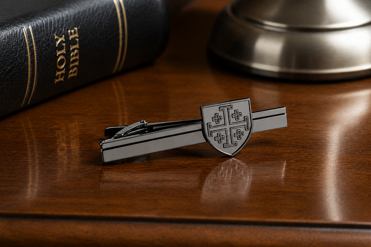A silver-toned metal tie clip featuring a shield crest design, displayed on a polished wooden desk next to a black leather Holy Bible and a blurred metallic object. Gunmetal Jerusalem Cross shield tie clip Christian tie bar for men