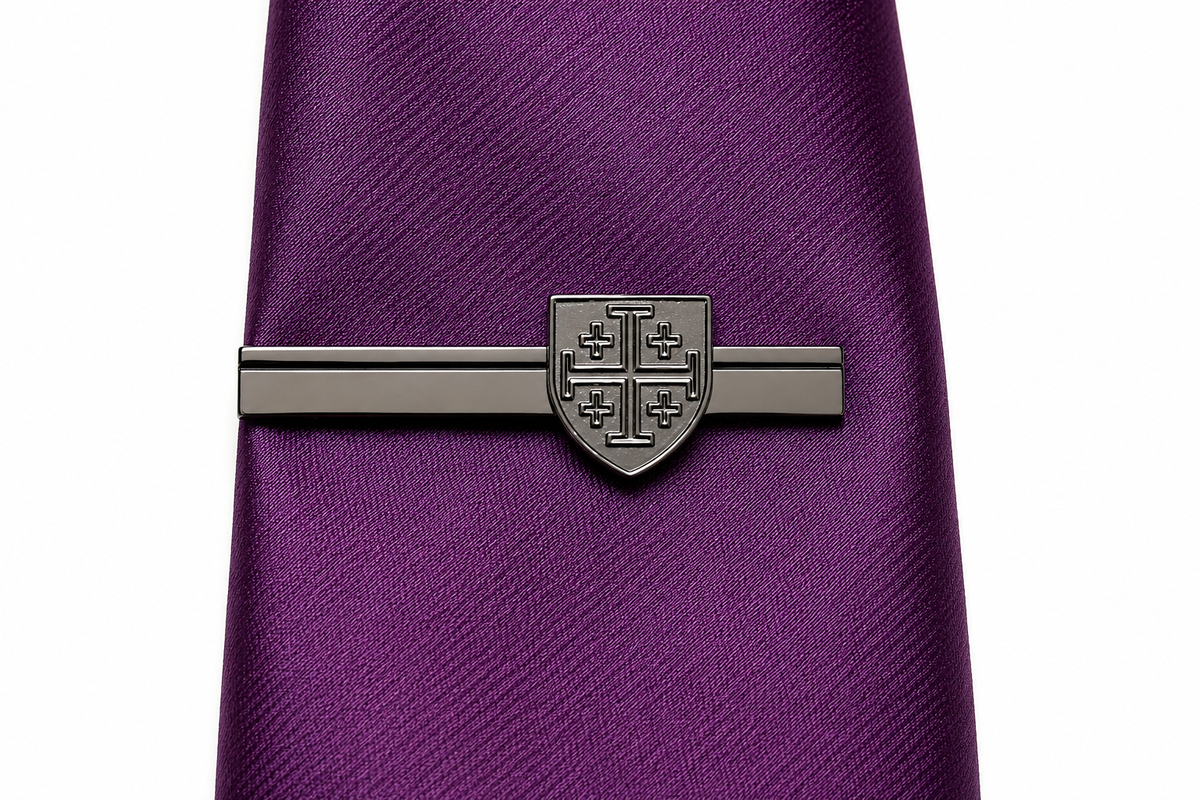 A close-up of a purple textured necktie with a dark metallic tie clip featuring a shield crest design, set against a plain white background. Gunmetal Jerusalem Cross shield tie clip Christian tie bar for men