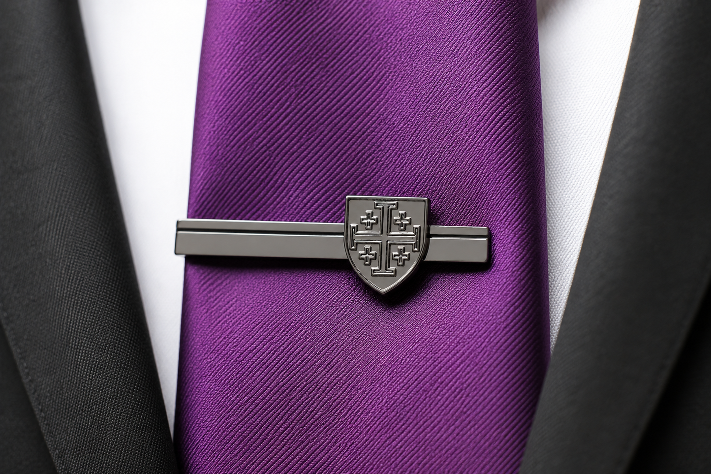 A dark gray metal tie clip featuring a shield crest design, shown fastening a purple textured necktie on a white dress shirt, framed by black suit jacket lapels.