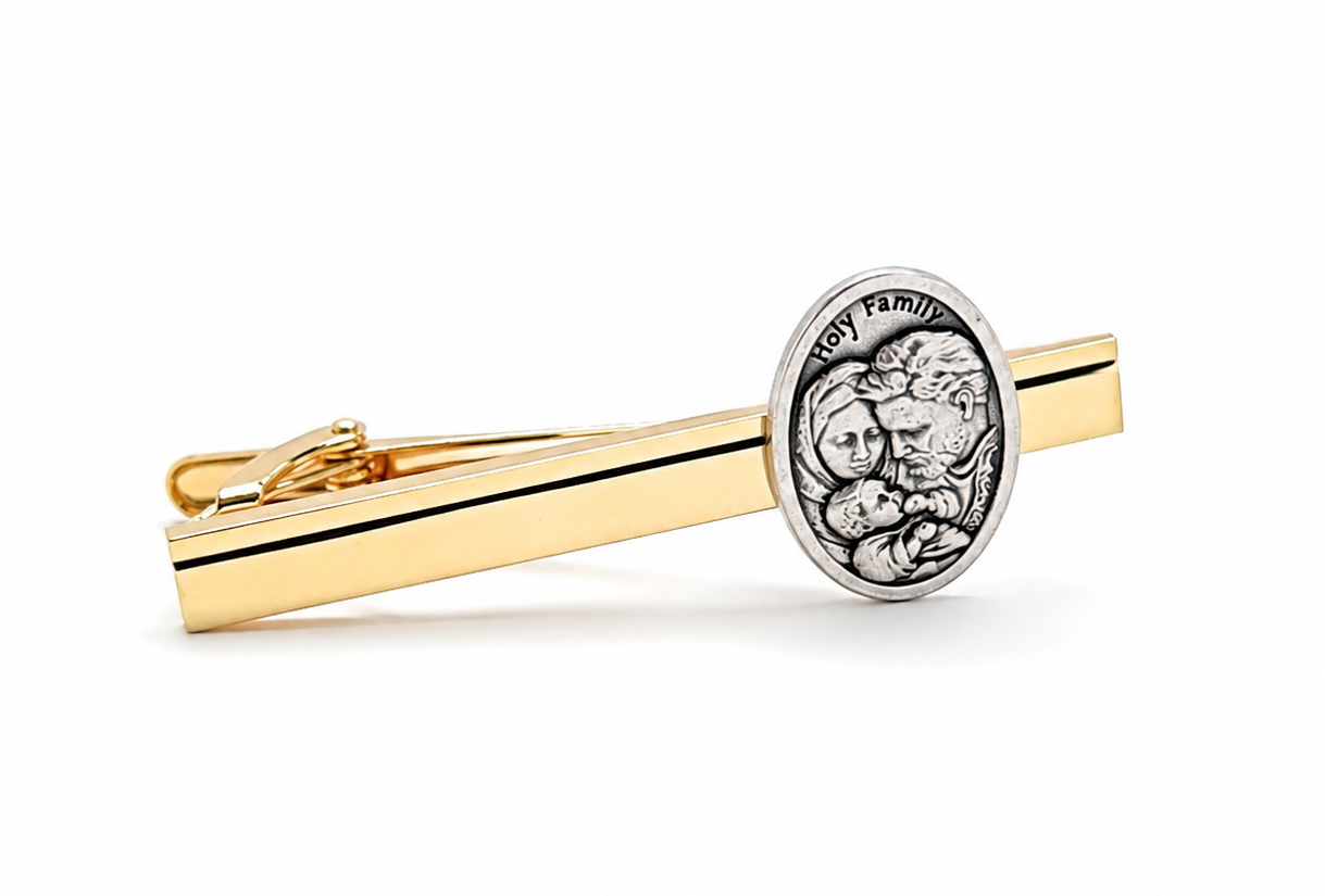 A gold and silver tie clip featuring a polished gold bar with a thin black stripe and a round silver-toned medallion engraved with a religious figure and the text Holy family