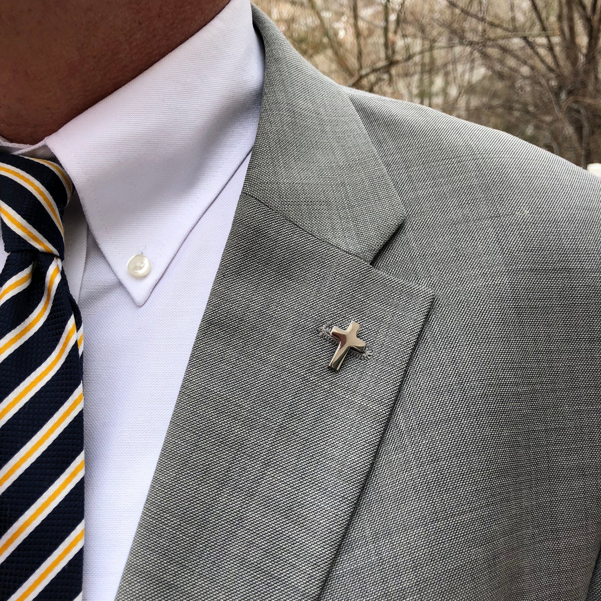 A silver-colored Christian cross lapel pin with a butterfly back, displayed against a white background. Gray suite background