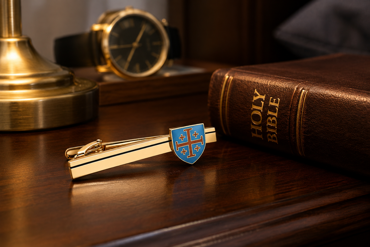 A gold-tone tie clip featuring a shield emblem with a red cross on a blue background, resting on a dark wooden desk next to a brown leather Holy Bible and a gold watch.
