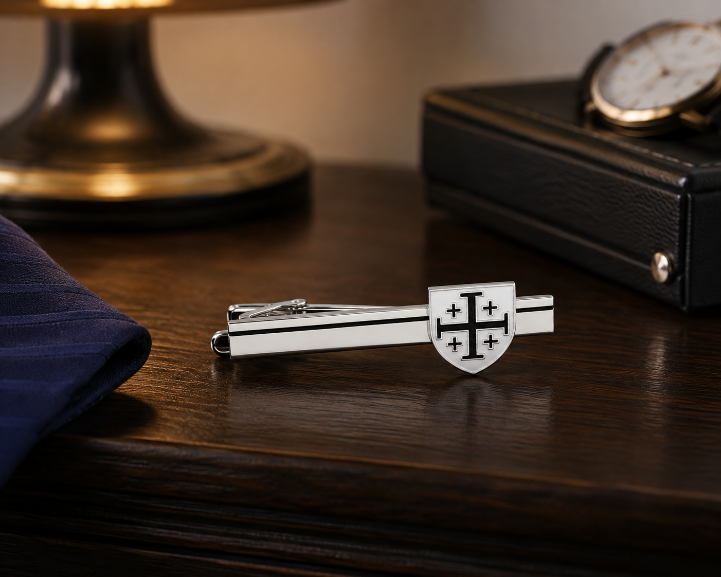 A silver-toned tie clip with a shield emblem featuring a black cross, displayed on a dark wooden table next to a folded navy blue tie, with a blurred watch and lamp in the background.