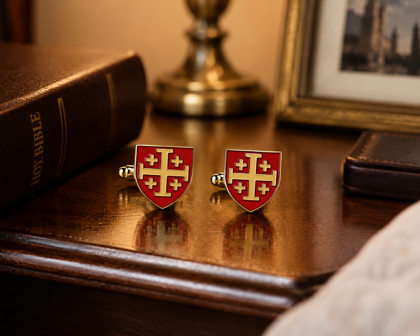 A pair of red and gold shield-shaped cufflinks with a cross design, displayed on a polished wooden table next to a leather-bound book.