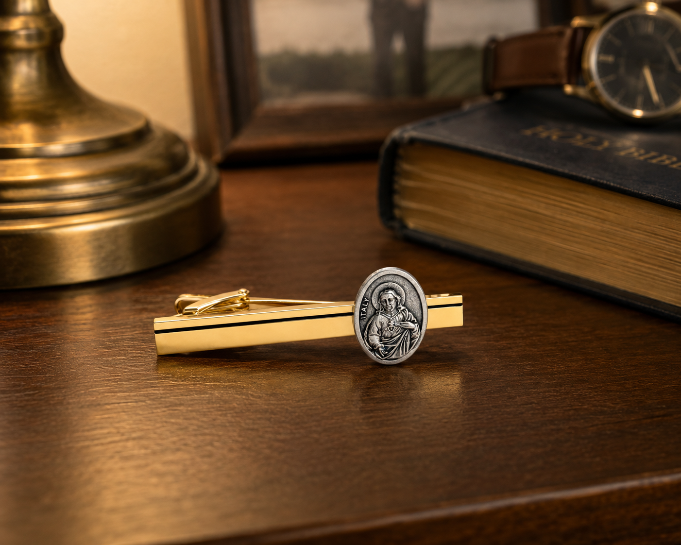 A gold and silver tie clip resting on a dark wooden table, styled with a vintage book, a watch, and a brass lamp base in the background.