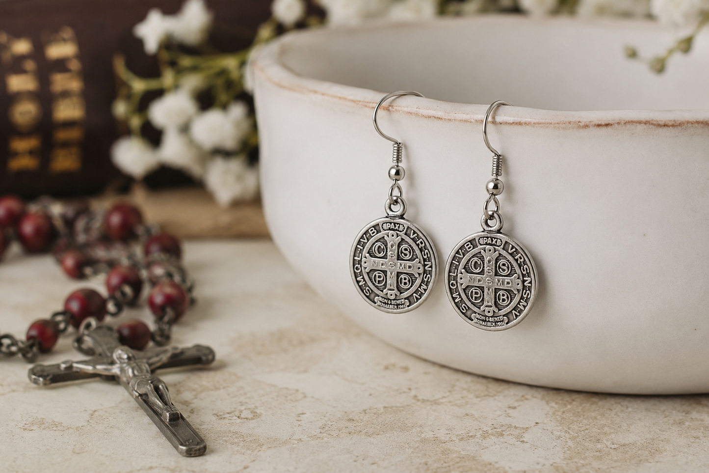 Silver earrings with circular designs on a rustic surface with a bowl and rosary in the background. Saint Benedict Medal Earrings Silver Catholic Christian Dangle Earrings for Women