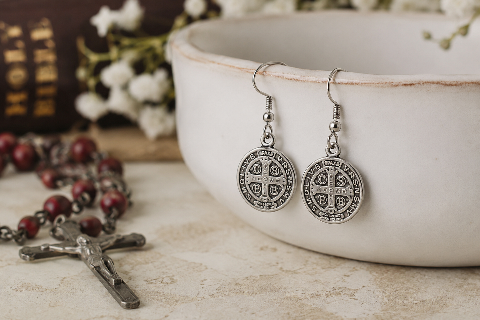 Silver earrings with circular designs on a rustic surface with a bowl and rosary in the background. Saint Benedict Medal Earrings Silver Catholic Christian Dangle Earrings for Women