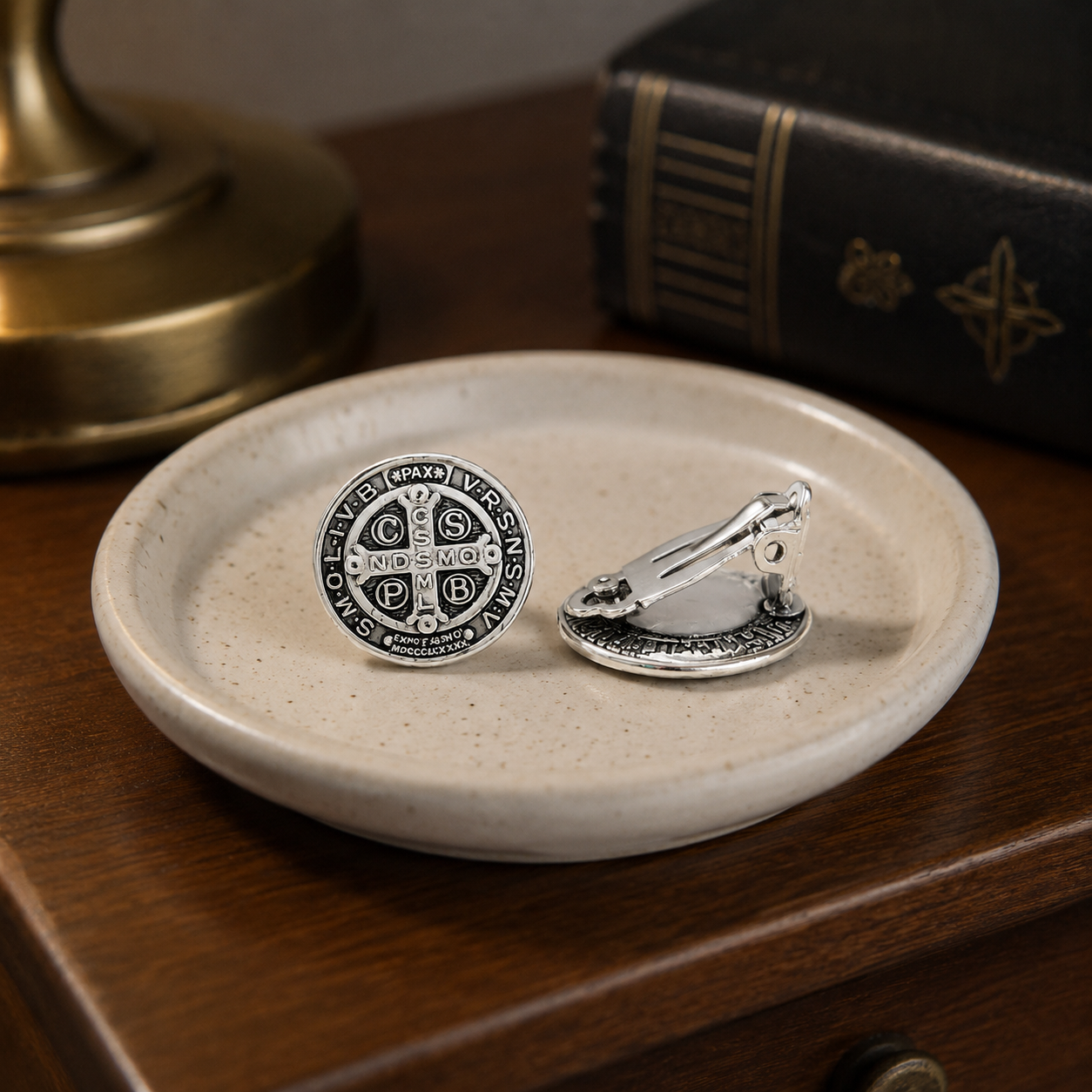 Silver Saint Benedict clip-on earrings displayed in a ceramic jewelry dish on a wooden nightstand.