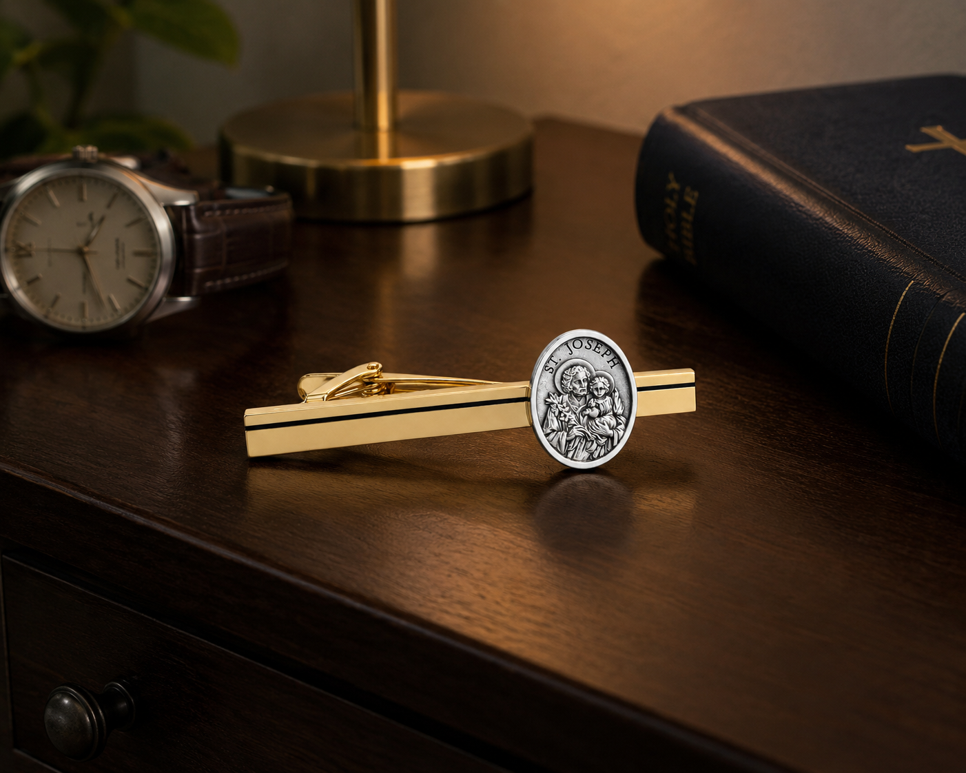A gold tie bar with a silver Saint Joseph coin emblem, displayed on a dark wooden desk alongside a watch, a brass lamp base, and a book.