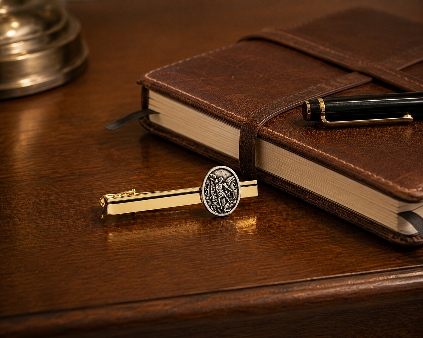 A gold and silver tie clip with an engraved circular emblem rests on a polished dark wood desk next to a brown leather notebook and a black pen.