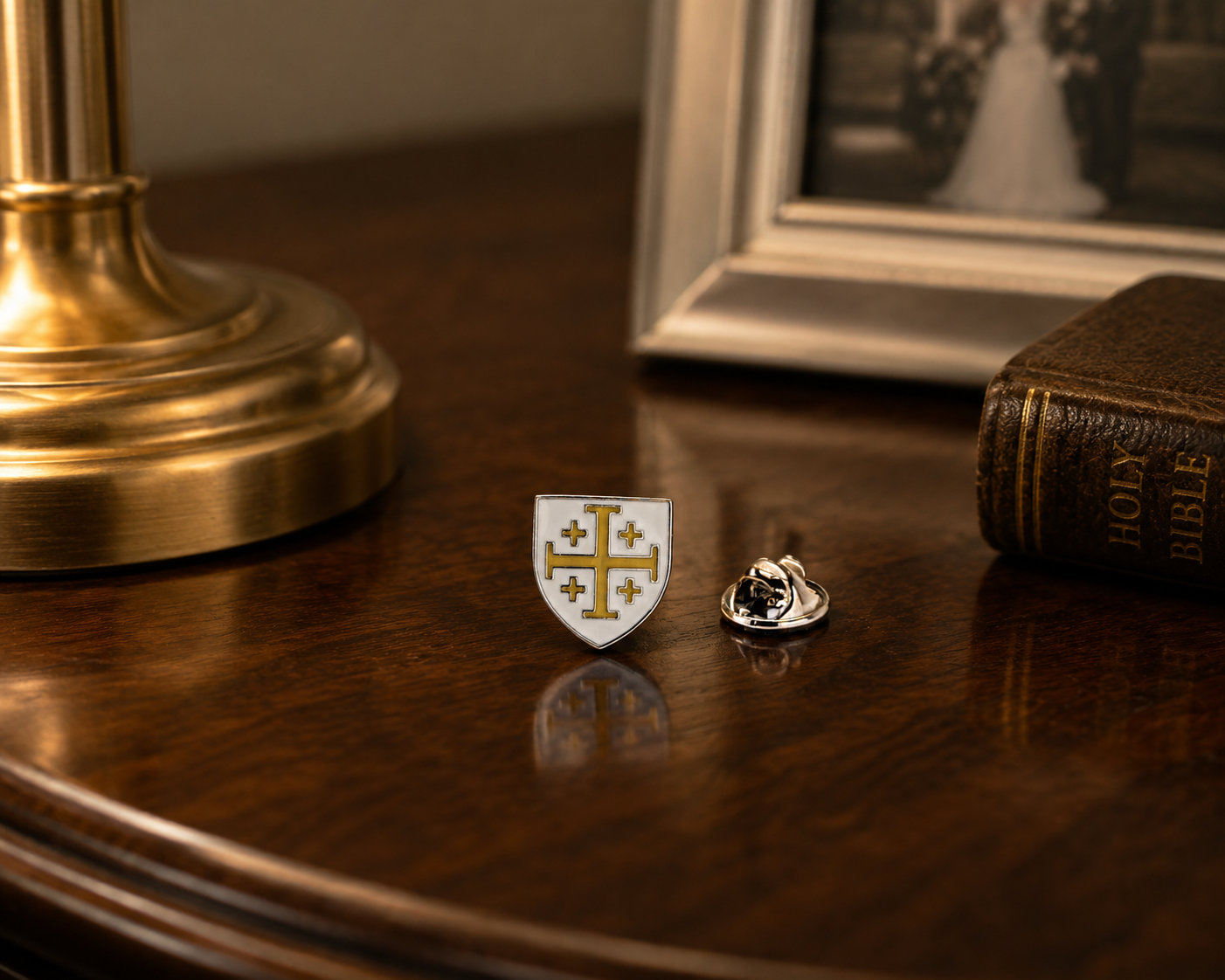 A small white shield-shaped pin with a gold cross and four stars, shown next to a silver-toned locket on a polished dark wood table, with a brass lamp base and a vintage Bible in the background.