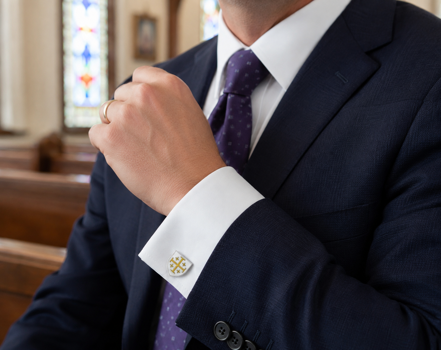 A close-up of a man wearing a navy blue suit, white dress shirt, and a purple patterned tie, adjusting his collar. A gold cufflink with a cross design is visible on his white cuff, and a gold ring is on his finger.